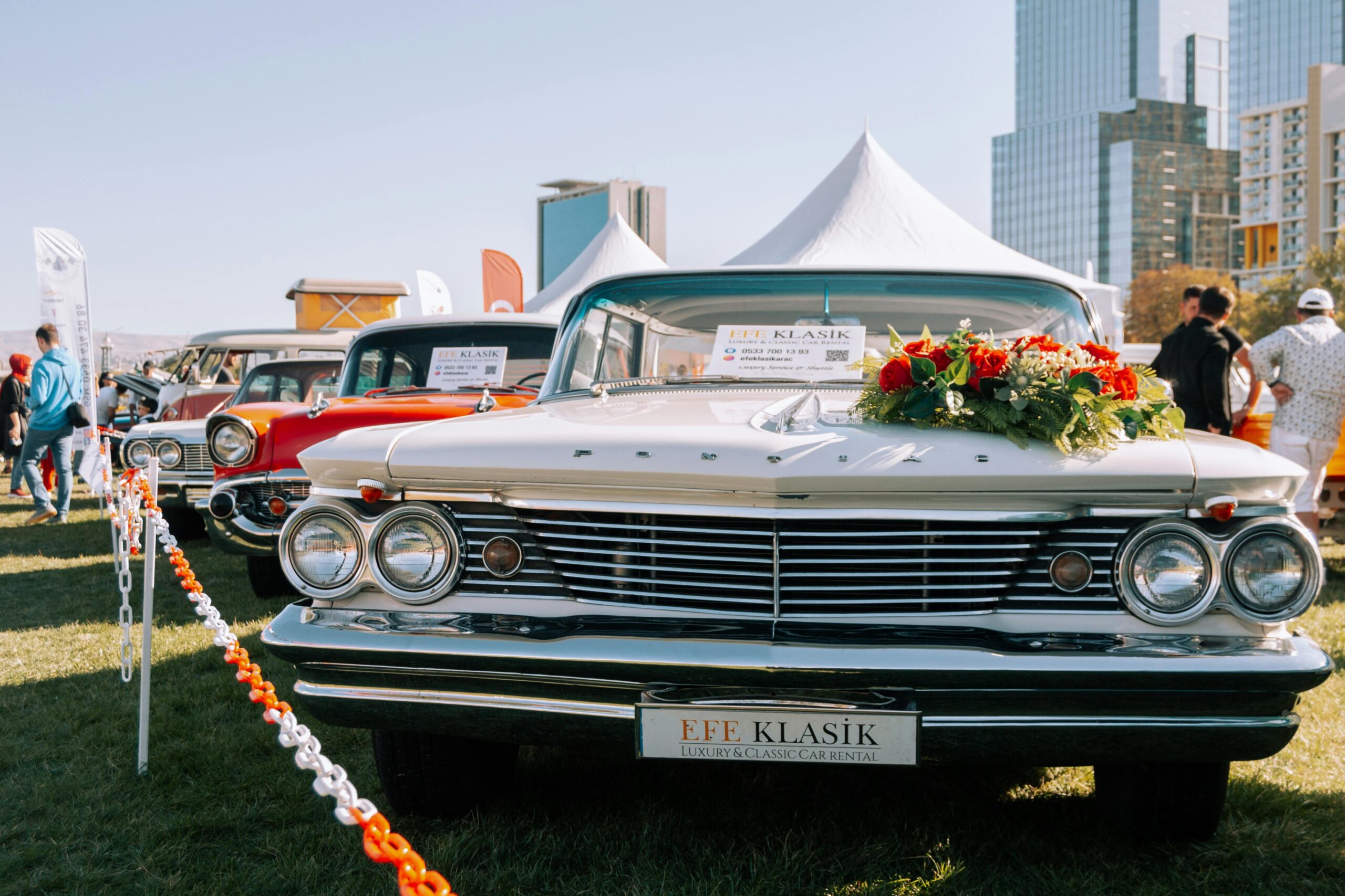 Front view of a classic white Pontiac with floral decorations at a car exhibition in a city setting.